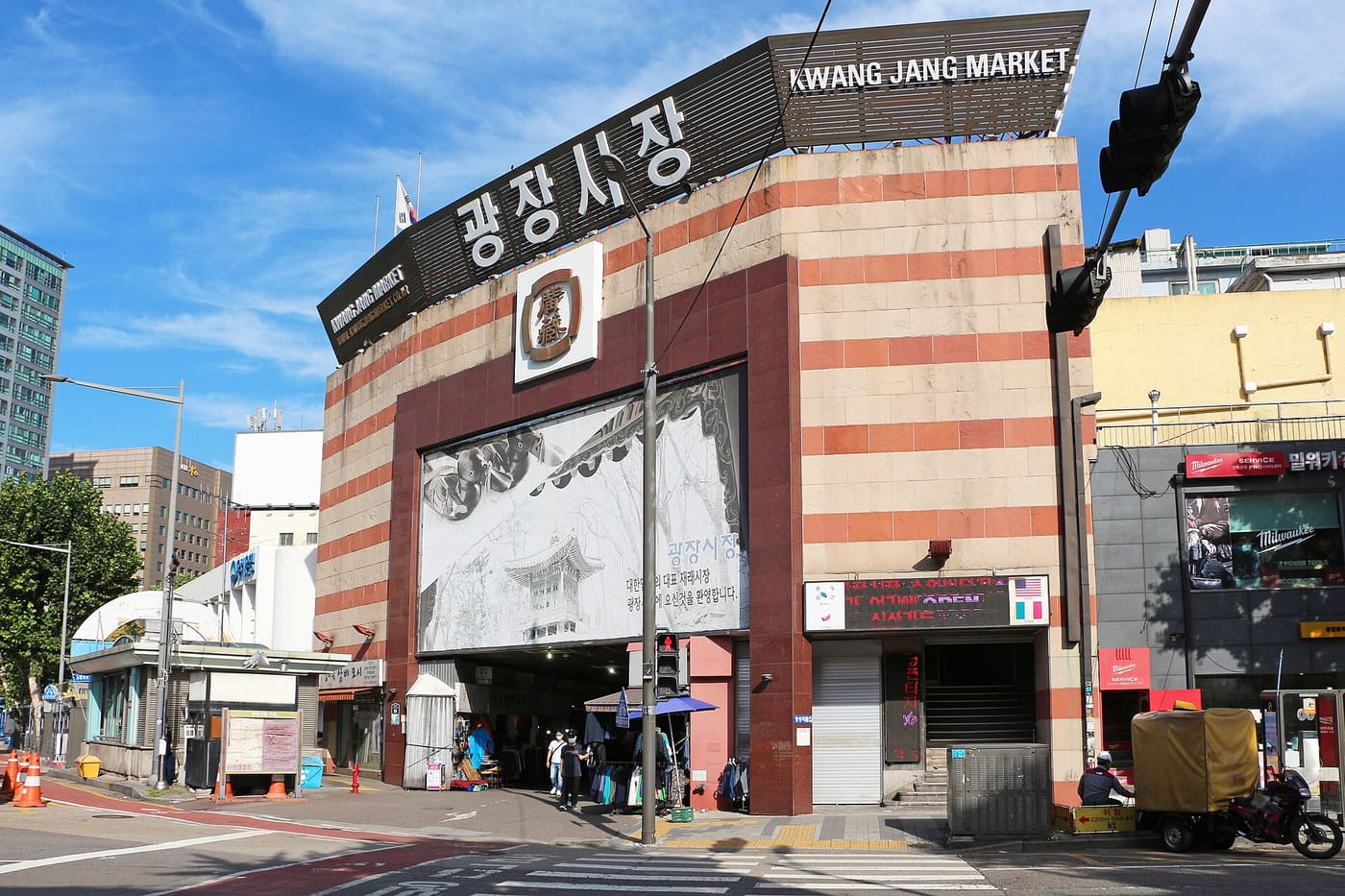 The exterior of Gwangjang Market in Seoul, a brick building with a large sign reading 광장시장 and KWANG JANG MARKET above an entrance arch, with the market visible through the opening.