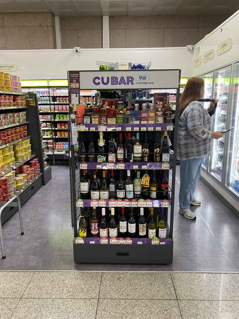 A wine rack inside a Korean CU convenience store, bottles arranged on shelves with price labels under a fixture marked CU BAR, while a customer stands at the drinks chiller behind it.