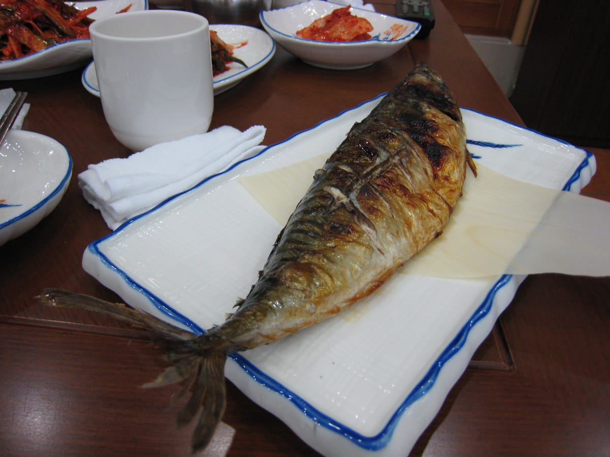 A whole grilled mackerel on a white plate at a Korean restaurant table, with kimchi and side dishes behind it.