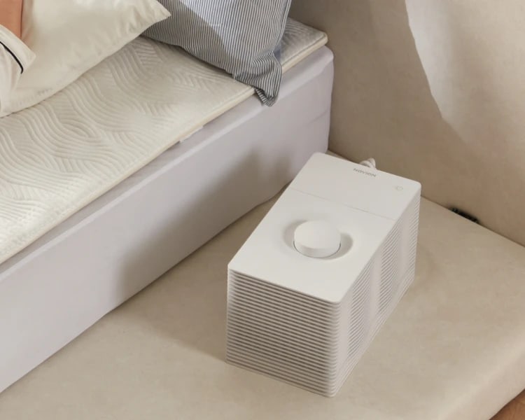 A Korean bedroom with a quilted heating mat on the mattress and a small white Navien boiler unit on the floor beside the bed, a control dial on its top face and a hose running from the back.