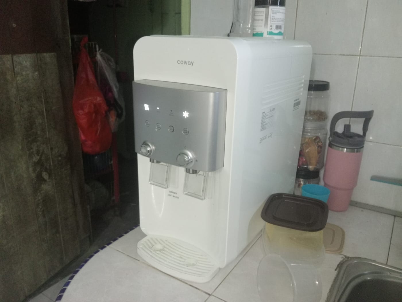 A white Coway water purifier with two dispensing taps on a kitchen counter in a lived-in home.