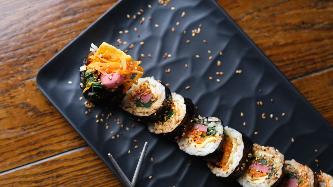 A row of Korean gimbap slices on a black plate with sesame seeds and metal chopsticks, showing spinach, ham, pickled radish, carrot and egg inside.