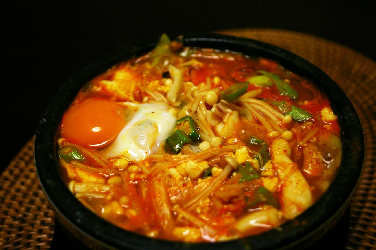 Sundubu-jjigae bubbling in a dark pot with a raw egg on top, soft tofu, courgette and enoki mushrooms in red broth.