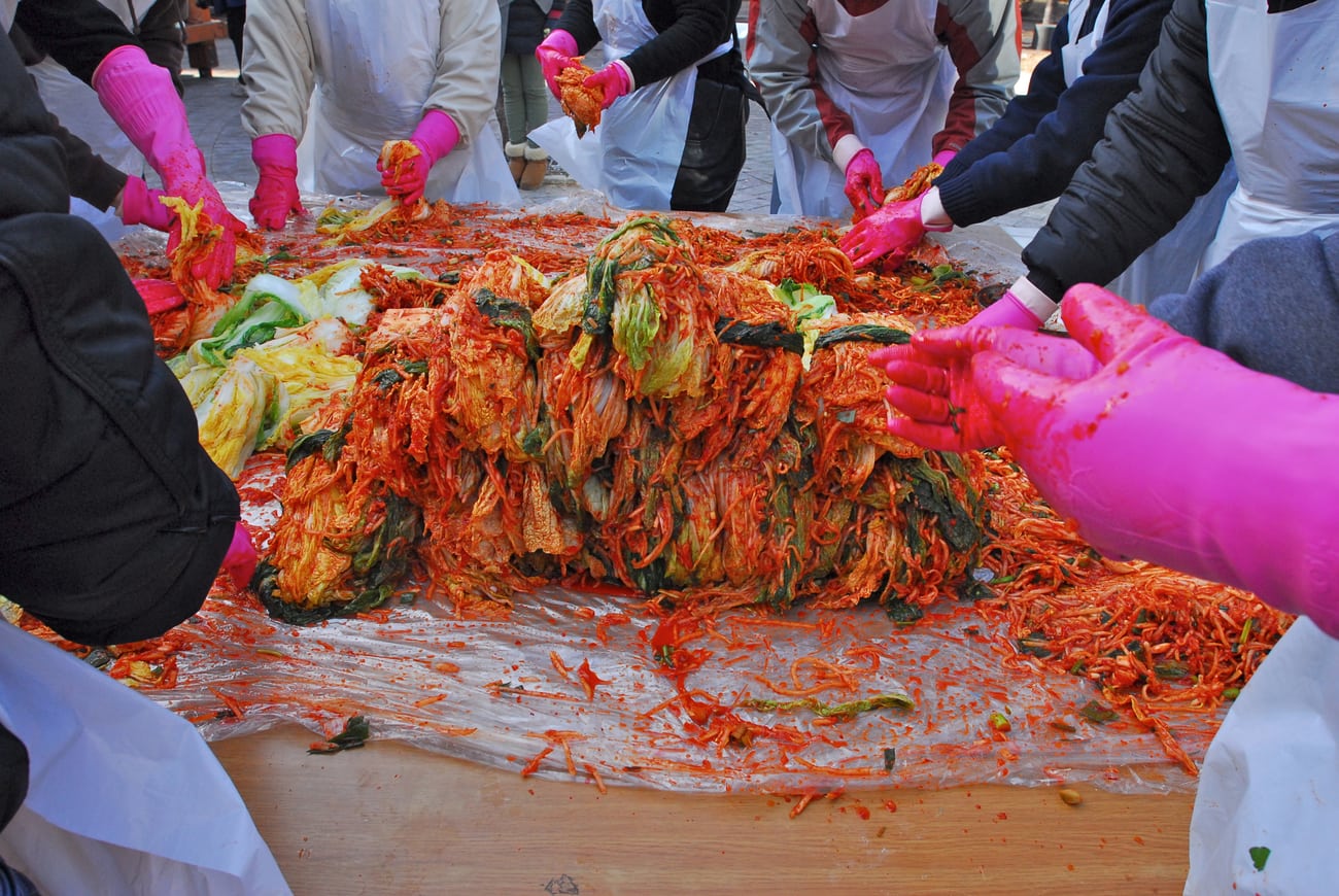 Volunteers in aprons and pink gloves making kimchi together at a long table piled with salted cabbage and red seasoning.