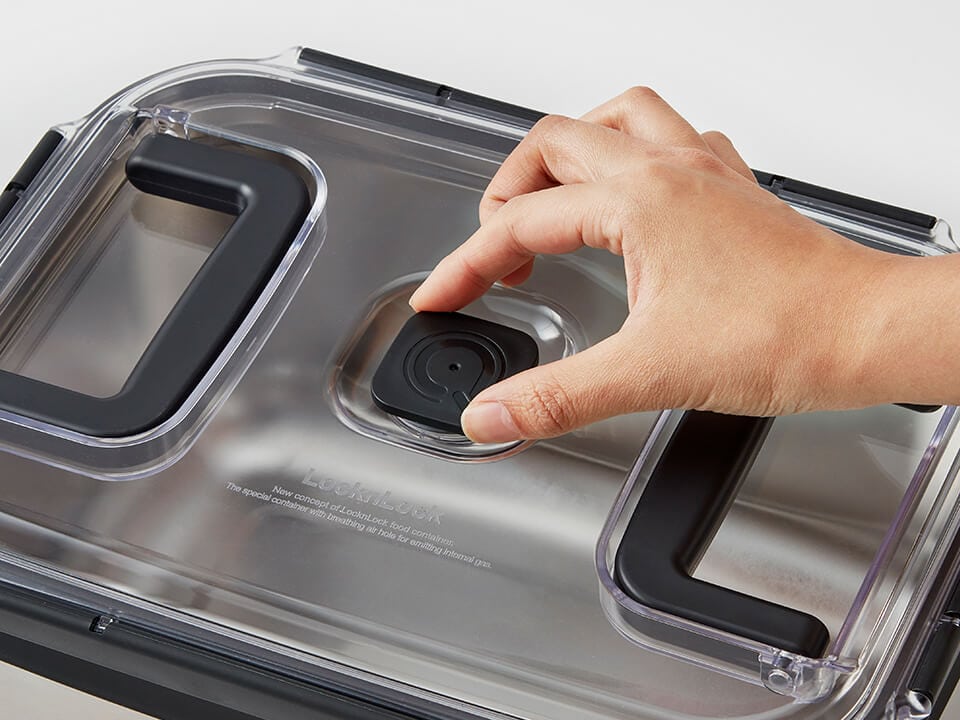 A hand pressing the round air valve on the clear lid of a stainless steel kimchi container.