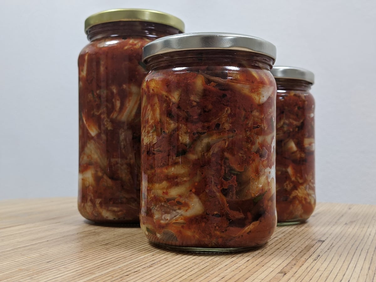 Three glass jars of kimchi with metal lids on a wooden surface.