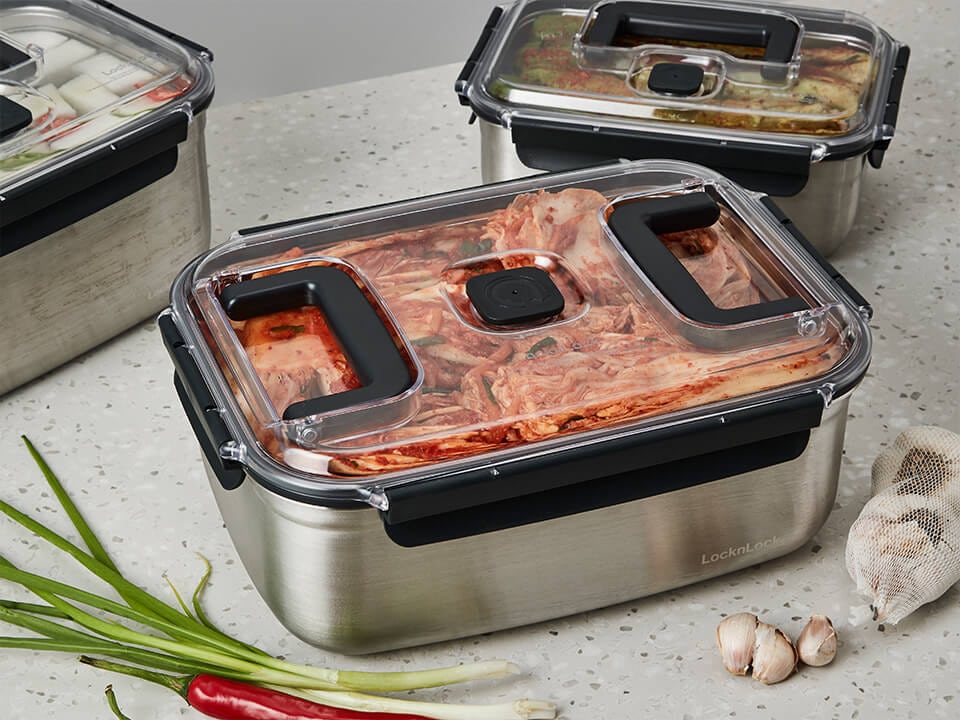 Rectangular stainless steel kimchi containers with clear lids, arranged side by side on a counter with spring onions and garlic.