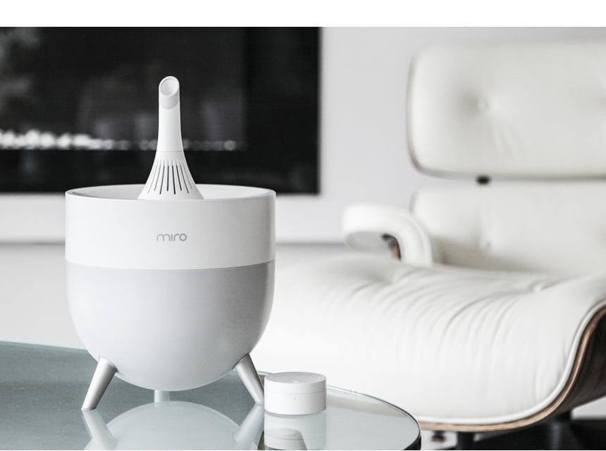 A white Miro humidifier standing on a glass side table in a bright living room, with a white leather lounge chair behind it and its small round refill cap resting beside it.