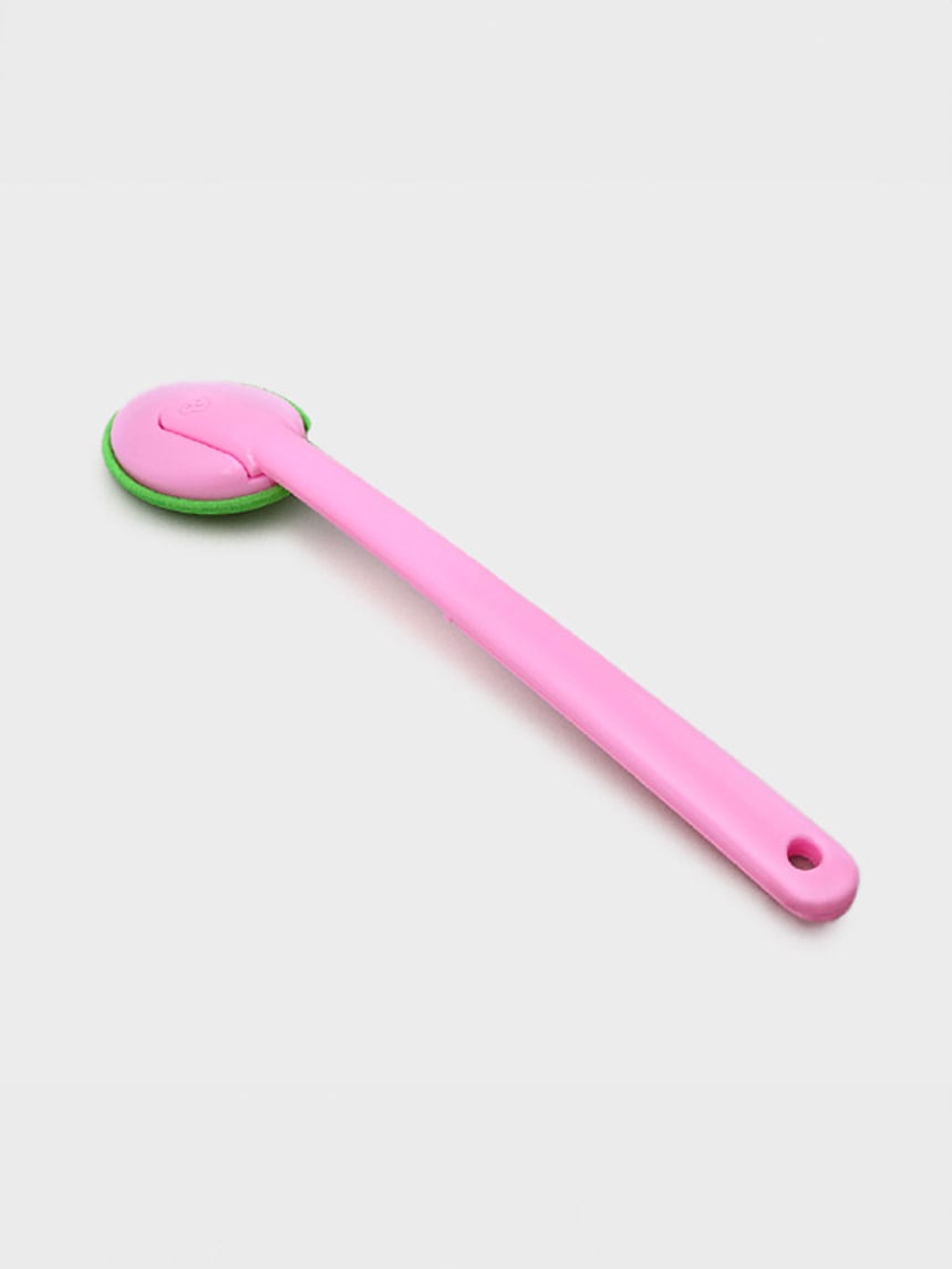 A pink plastic handled back scrubber with a round green exfoliating head, photographed against a plain background.