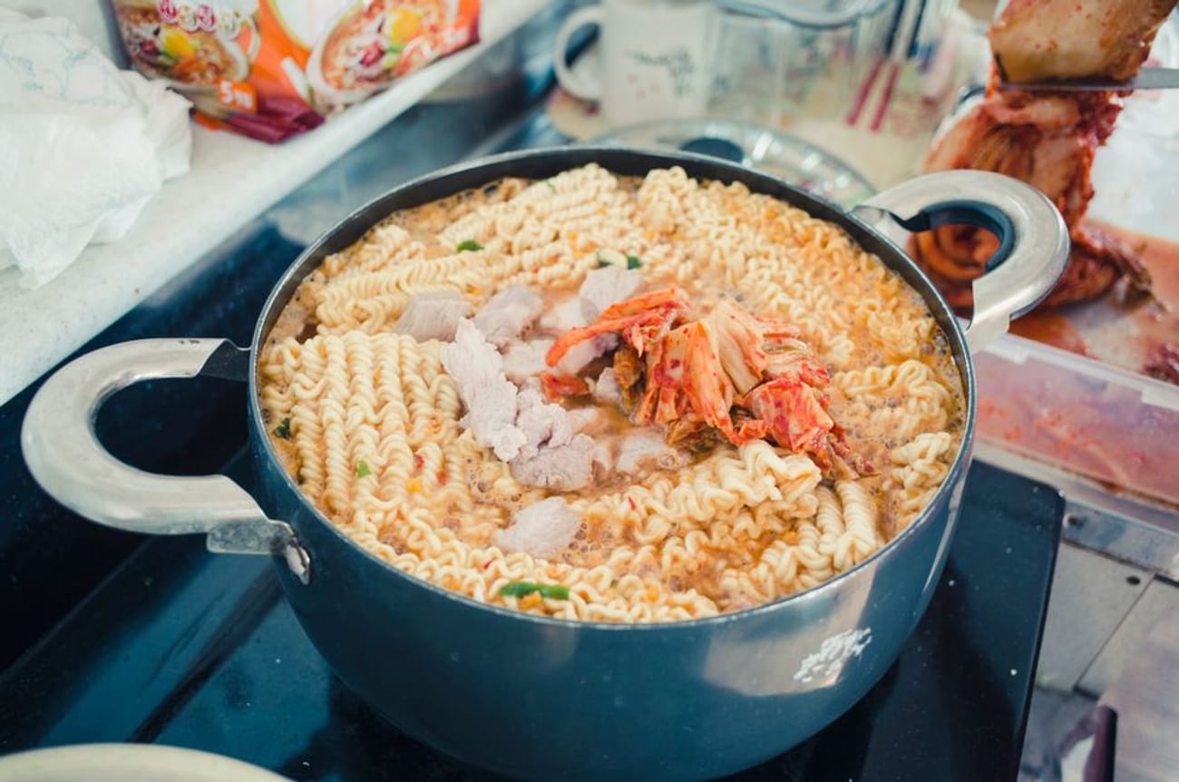Kimchi ramyun simmering in a metal pot on a stovetop, wavy noodles and kimchi visible in a red broth, with a Korean instant noodle packet in the background.