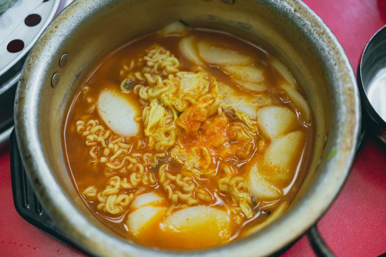 Rabokki — tteokbokki rice cakes cooked together with ramyun noodles in a red spicy broth — bubbling in a plain metal pot on a red table.