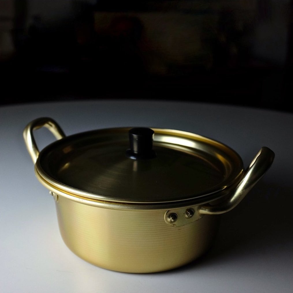 A gold anodised aluminium Korean ramyun pot with two gold side handles, a matching gold lid and a black knob, lit against a dark background.