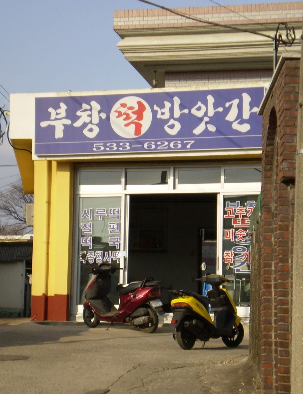 A small Korean neighbourhood mill on a village street, its blue sign reading 부창 떡방앗간 with a phone number, and hand-painted window text listing gochugaru, misutgaru and chamgireum.