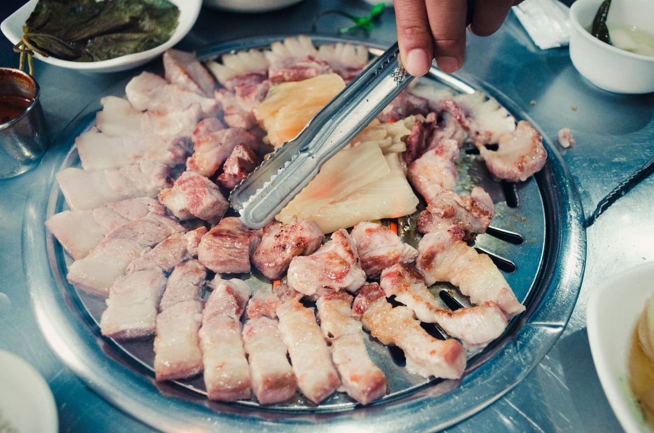 Slices of pork belly cooking on a round Korean grill plate, a hand turning a piece with metal tongs, with lettuce and side dishes around the edge of the table.
