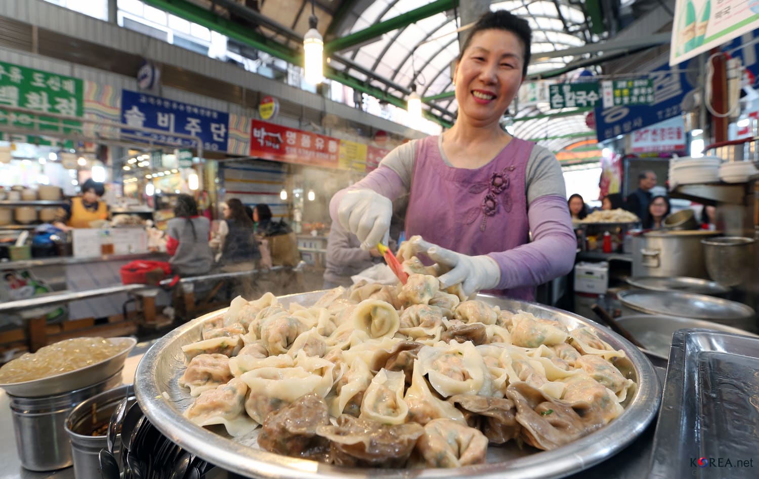 A smiling vendor in a purple apron at Gwangjang Market, Seoul, steaming a large tray of Korean dumplings under a hand-painted menu sign.