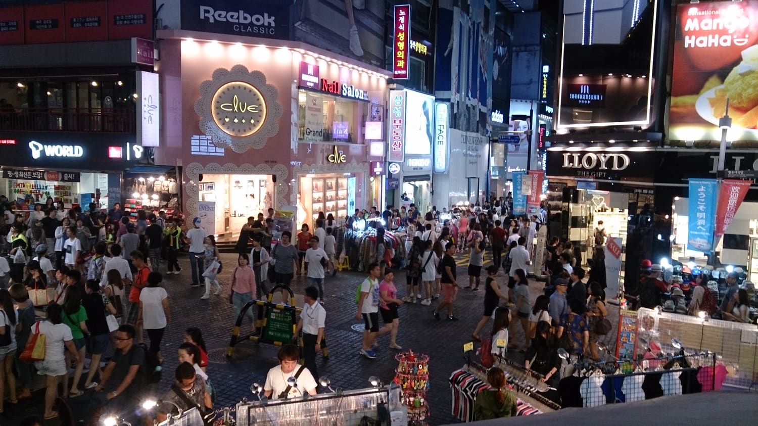 A dense night-time crowd of shoppers and tourists filling a Myeongdong street in Seoul, neon shop signs on both sides.