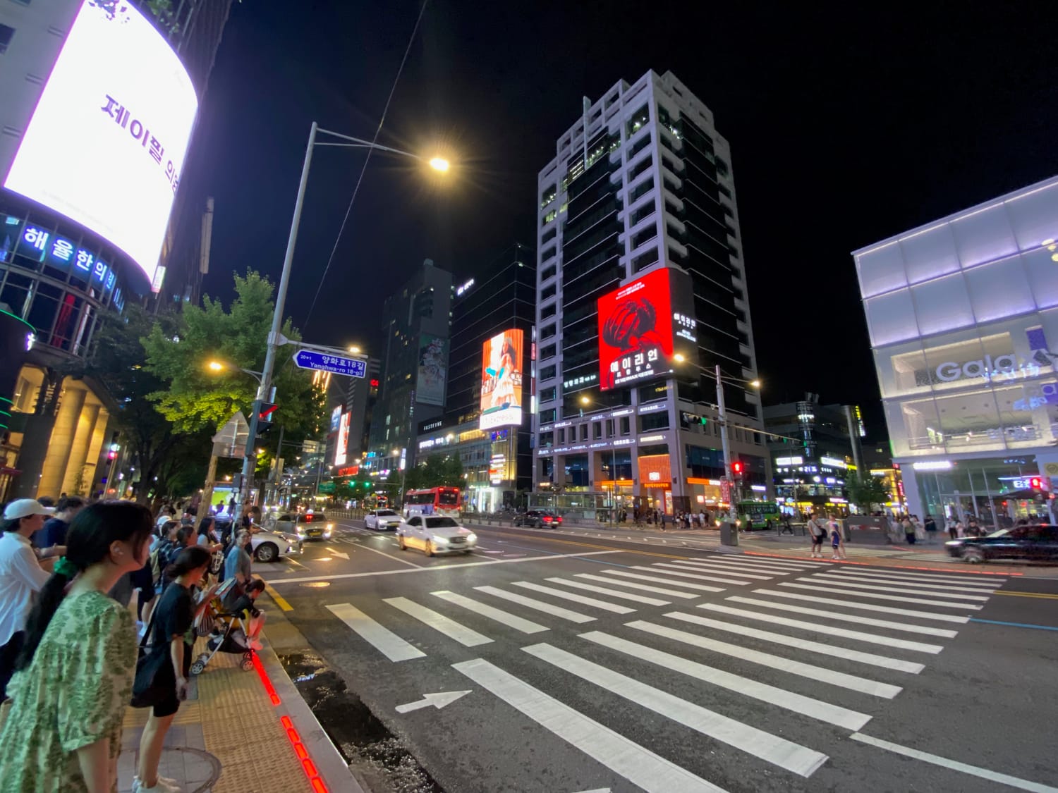 A night-time crossing in Hongdae, Seoul, with young people waiting at the lights under neon signs and a large screen.