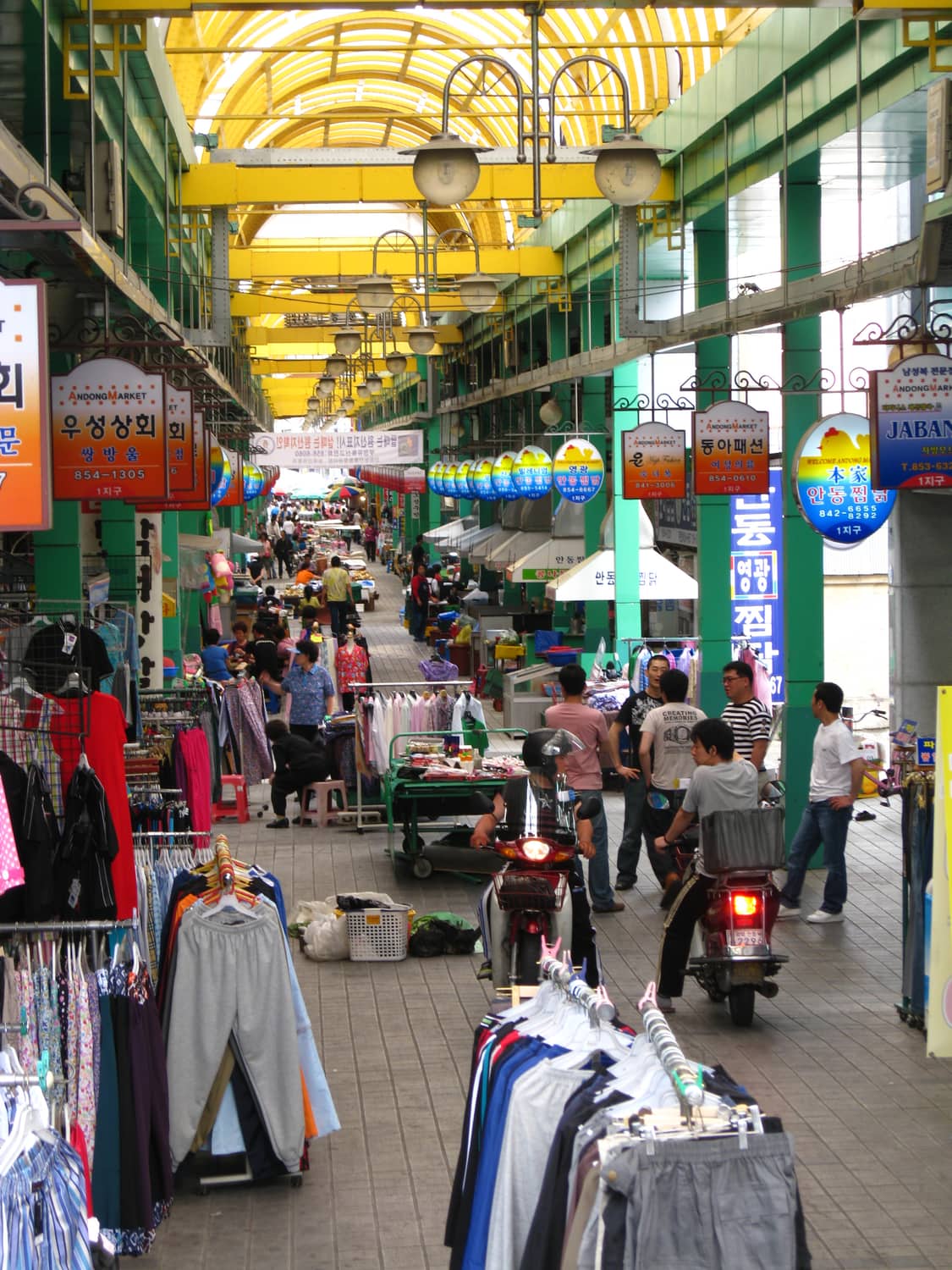 Inside a covered traditional market in Andong, shoppers and vendors under a high roof strung with signs.