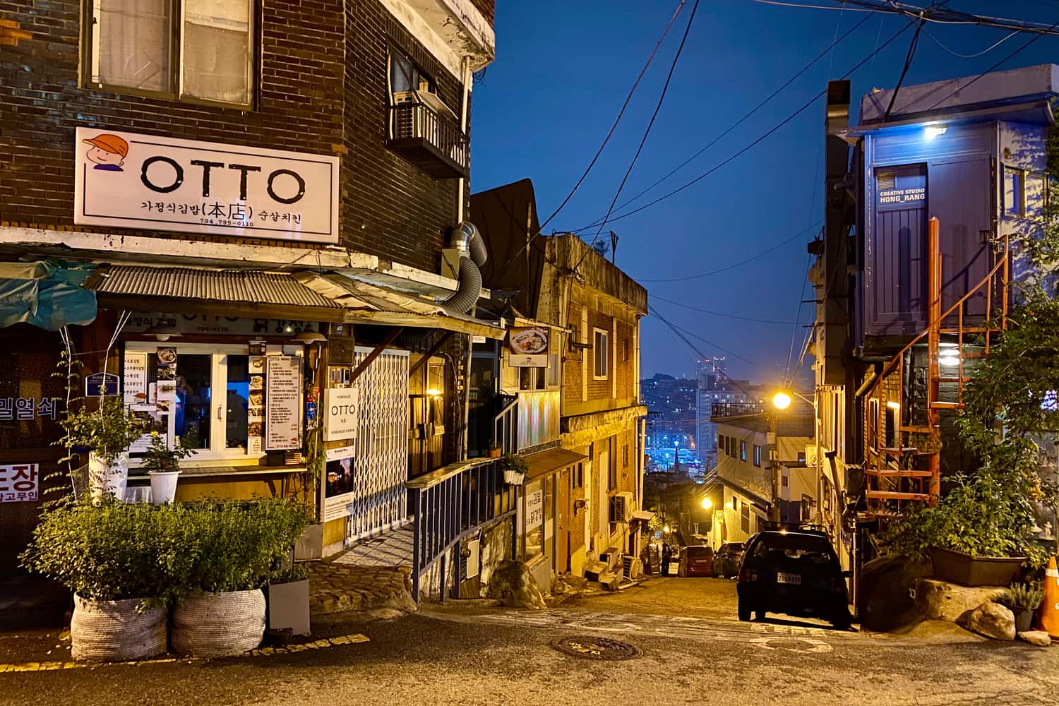 A street in Itaewon, Seoul, on a summer evening, low brick buildings with a shop sign reading OTTO and the hill rising behind.