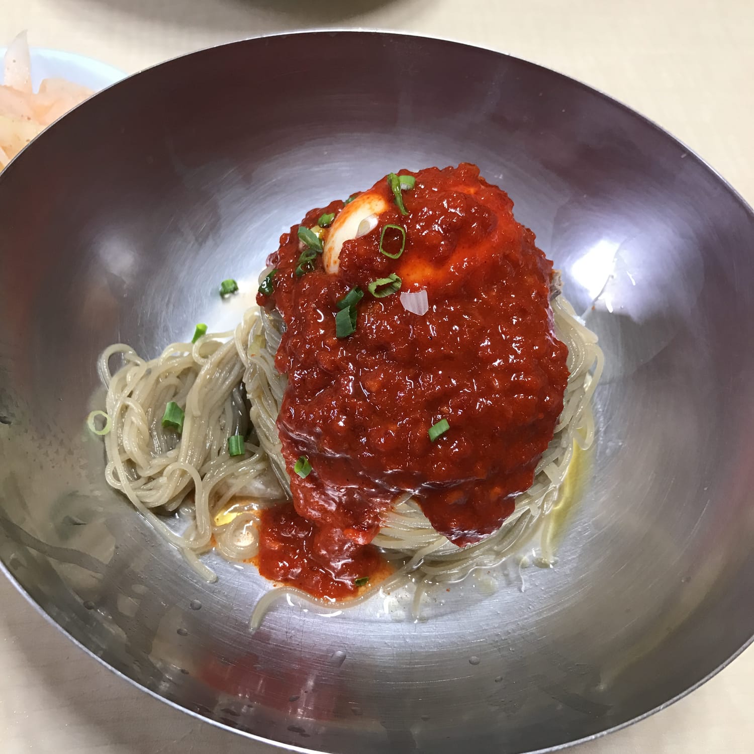 A stainless bowl of Hamheung-style bibim naengmyeon: translucent starch noodles under a glossy mound of red sauce, sliced egg and spring onion.