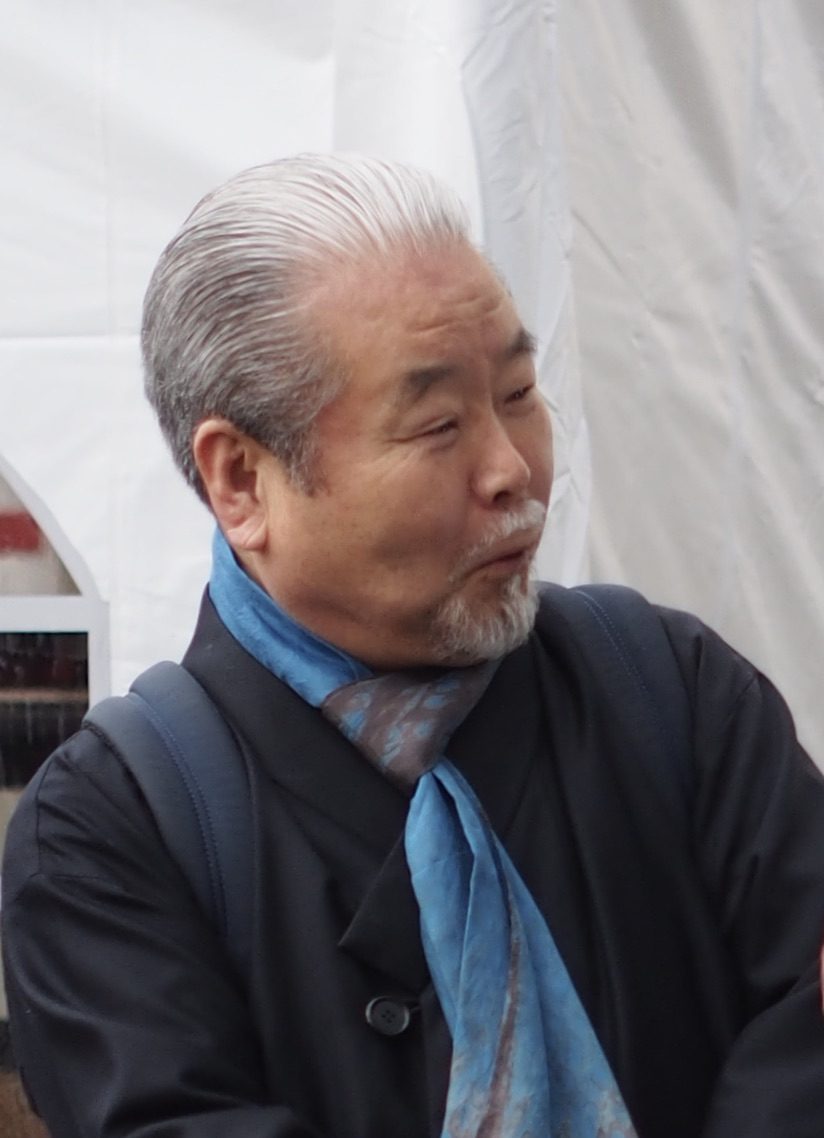 Kim Duk-soo, grey-haired with a goatee and a blue scarf, in profile at an outdoor celebration in Paris.