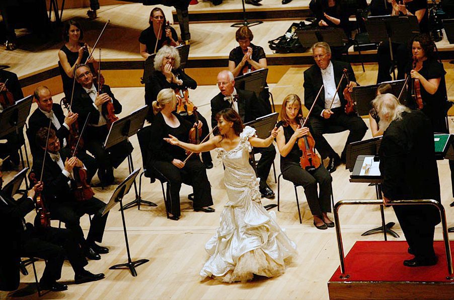 Sumi Jo in a white gown singing in front of an orchestra at a London concert hall, seen from above.