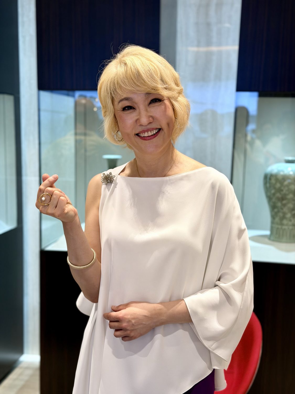 Jazz singer Youn Sun Nah, with short blonde hair and a white blouse, smiling and making a finger heart in front of a museum display case in Budapest.