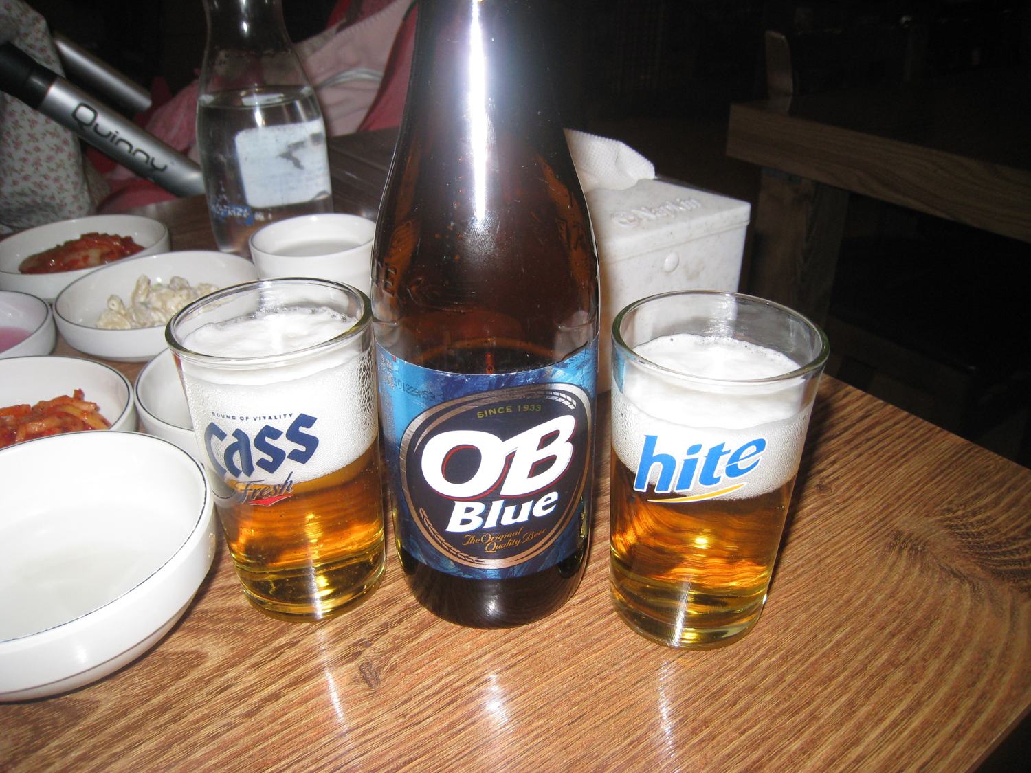 Three Korean beers on a restaurant table: a bottle of OB Blue between glasses of Cass and Hite, with side dishes.