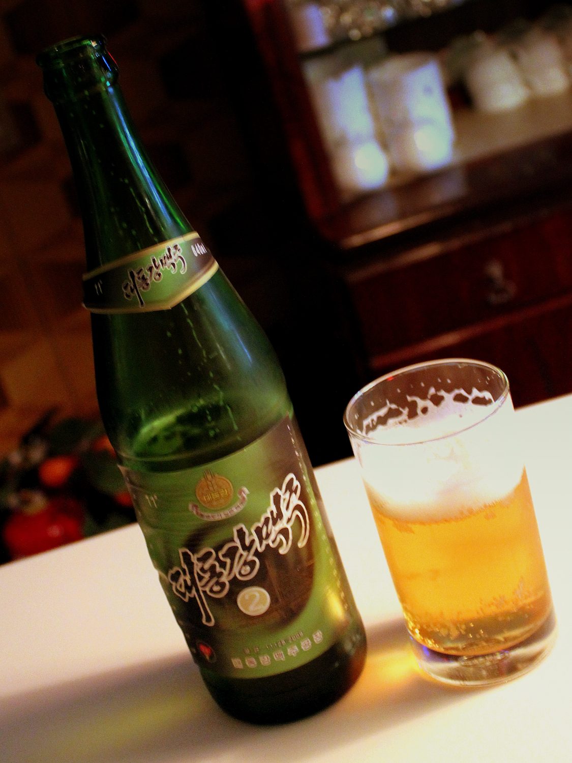 A green 640 ml bottle of Taedonggang beer beside a half-full glass on a bar counter in Pyongyang.