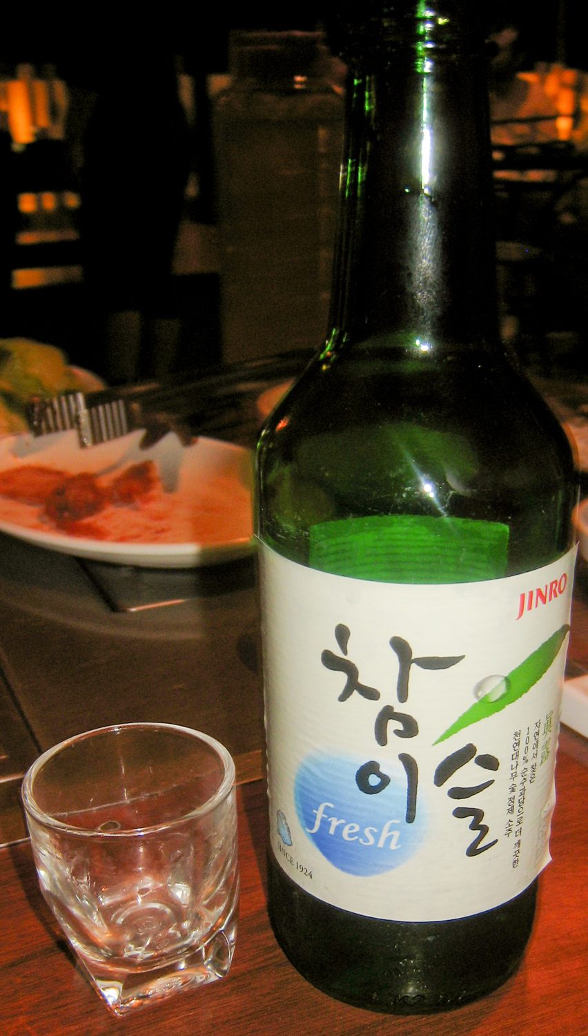 A bottle of Chamisul Fresh and a filled soju glass on a restaurant table beside plates of banchan.