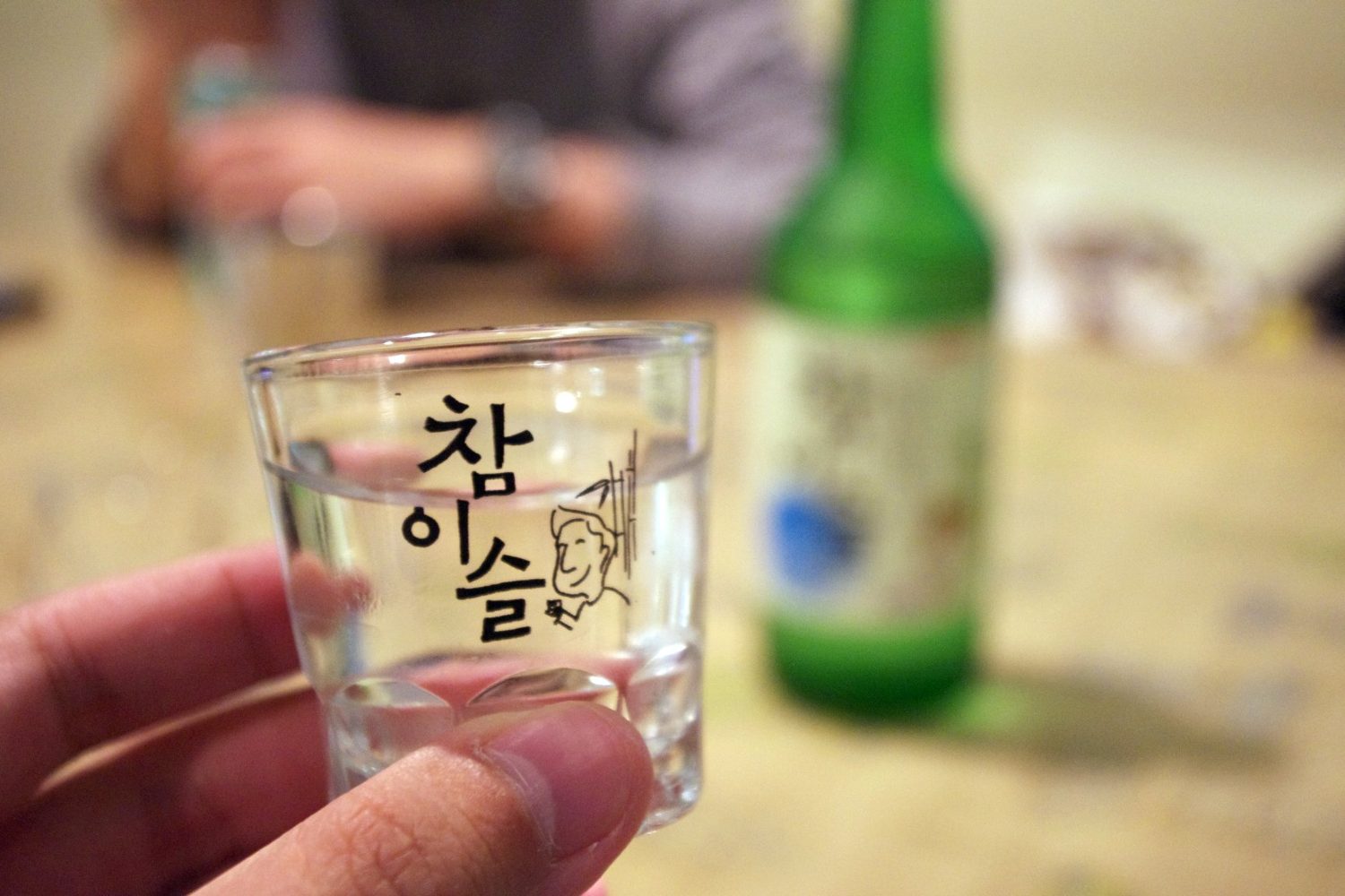 A hand holding a Chamisul-branded soju glass up to the camera across a restaurant table, a green bottle blurred behind.