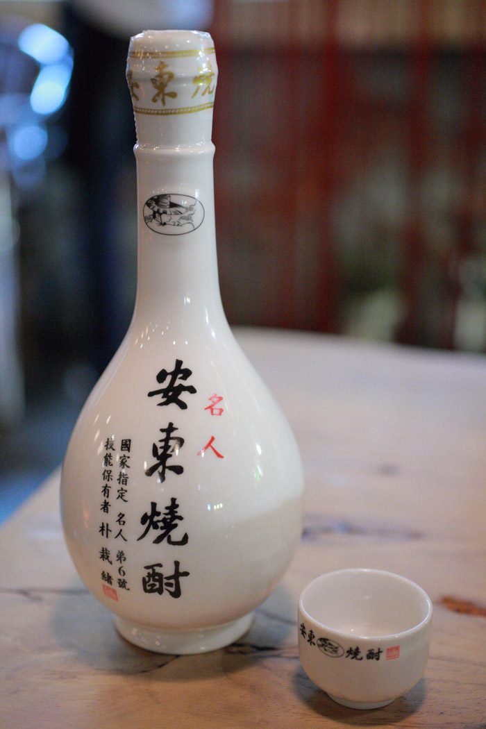 A single white gourd-shaped bottle of Park Jae-seo's Andong soju with a small cup, on a table.