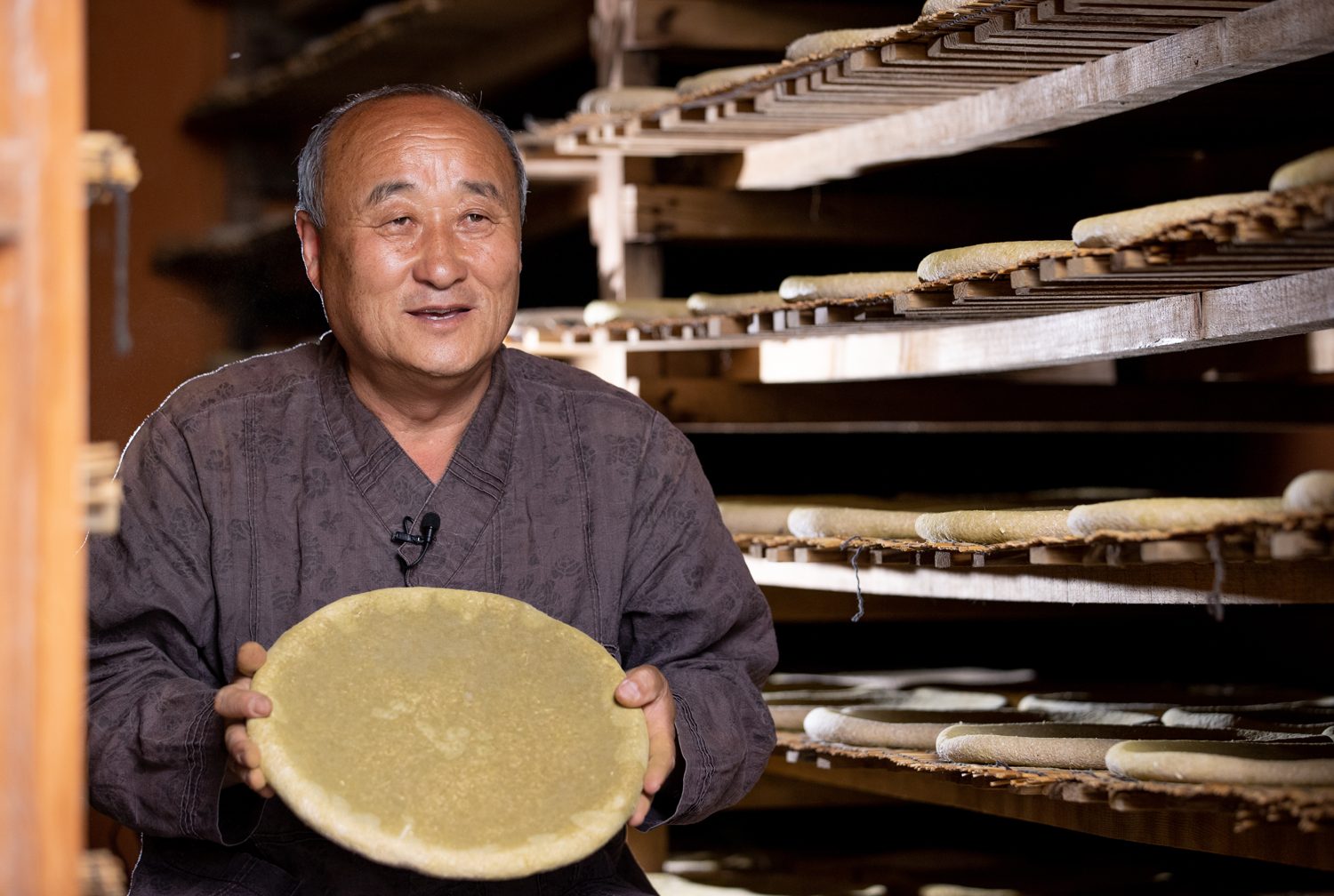 A brewer at Geumjeongsanseong, Busan, smiling and holding up a round disc of nuruk in front of drying racks.
