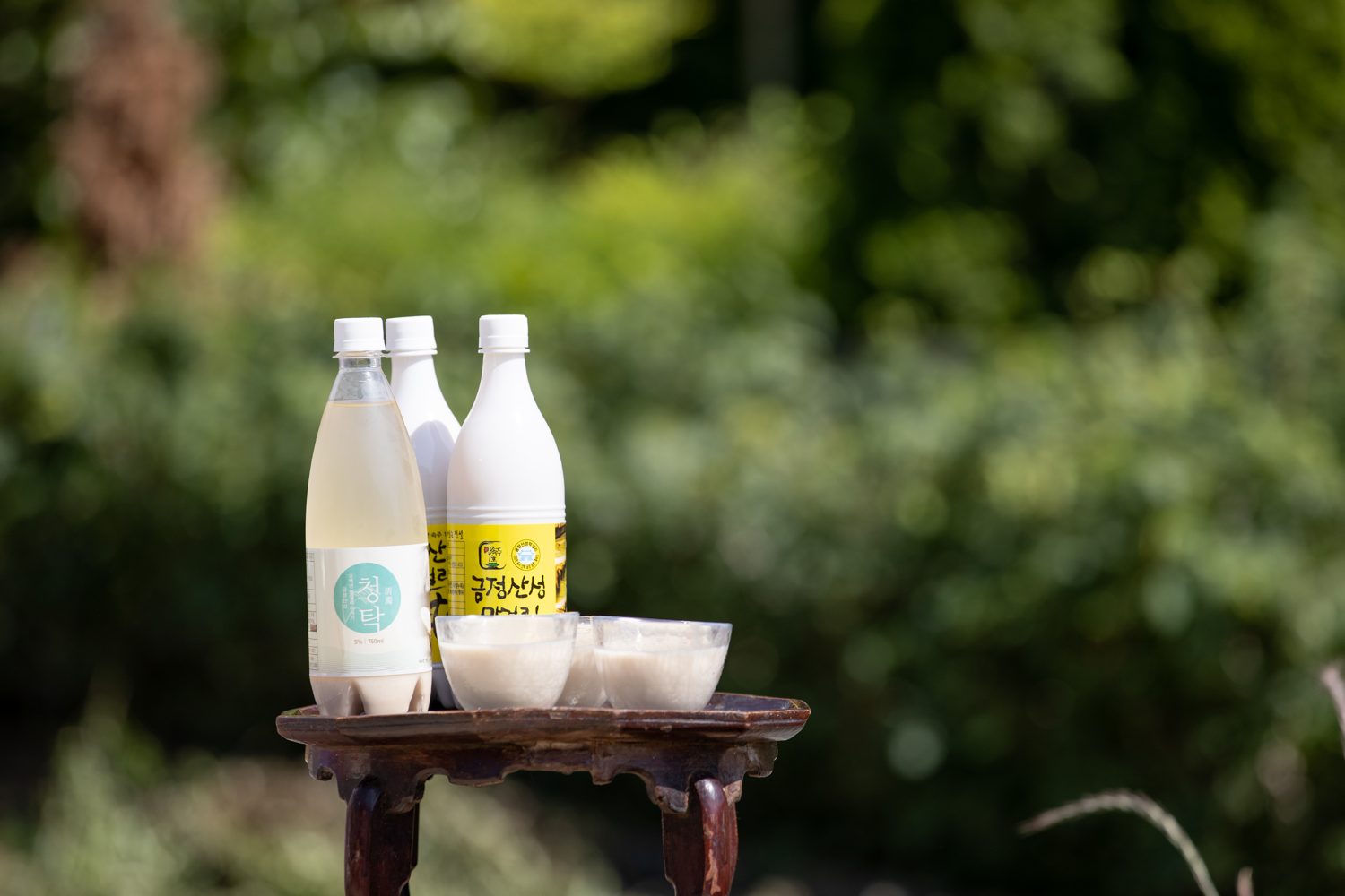 Three bottles of Geumjeongsanseong makgeolli and a set of small cups on a low wooden tray outdoors in Busan.
