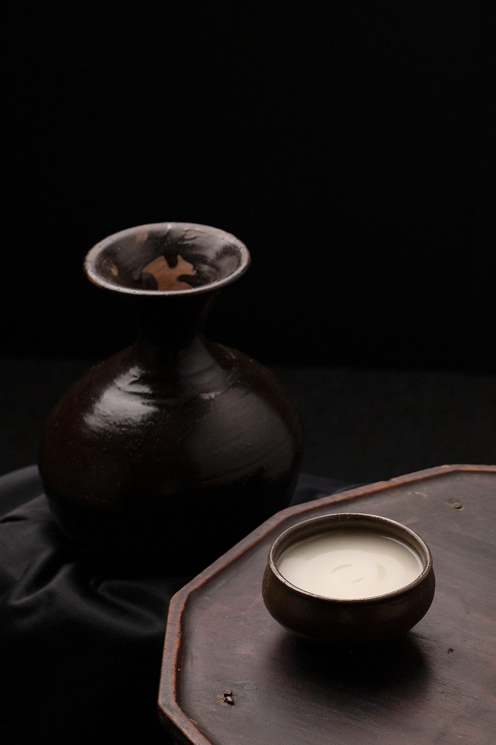 A brass makgeolli kettle and a bowl of makgeolli on a dark table.