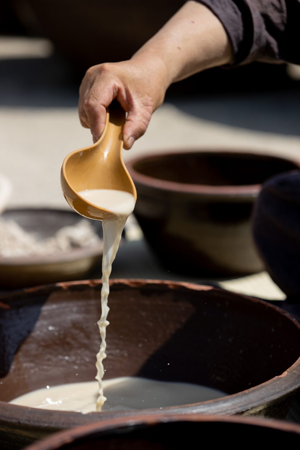 Makgeolli being poured from a gourd ladle into an earthenware jar, hands in traditional dress.