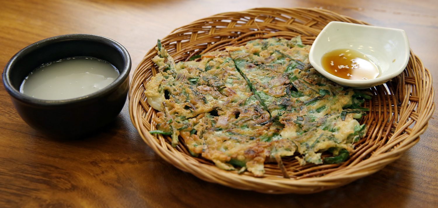 A basket of scallion pancake beside a dark ceramic bowl of makgeolli and a dipping sauce, on a wooden table.
