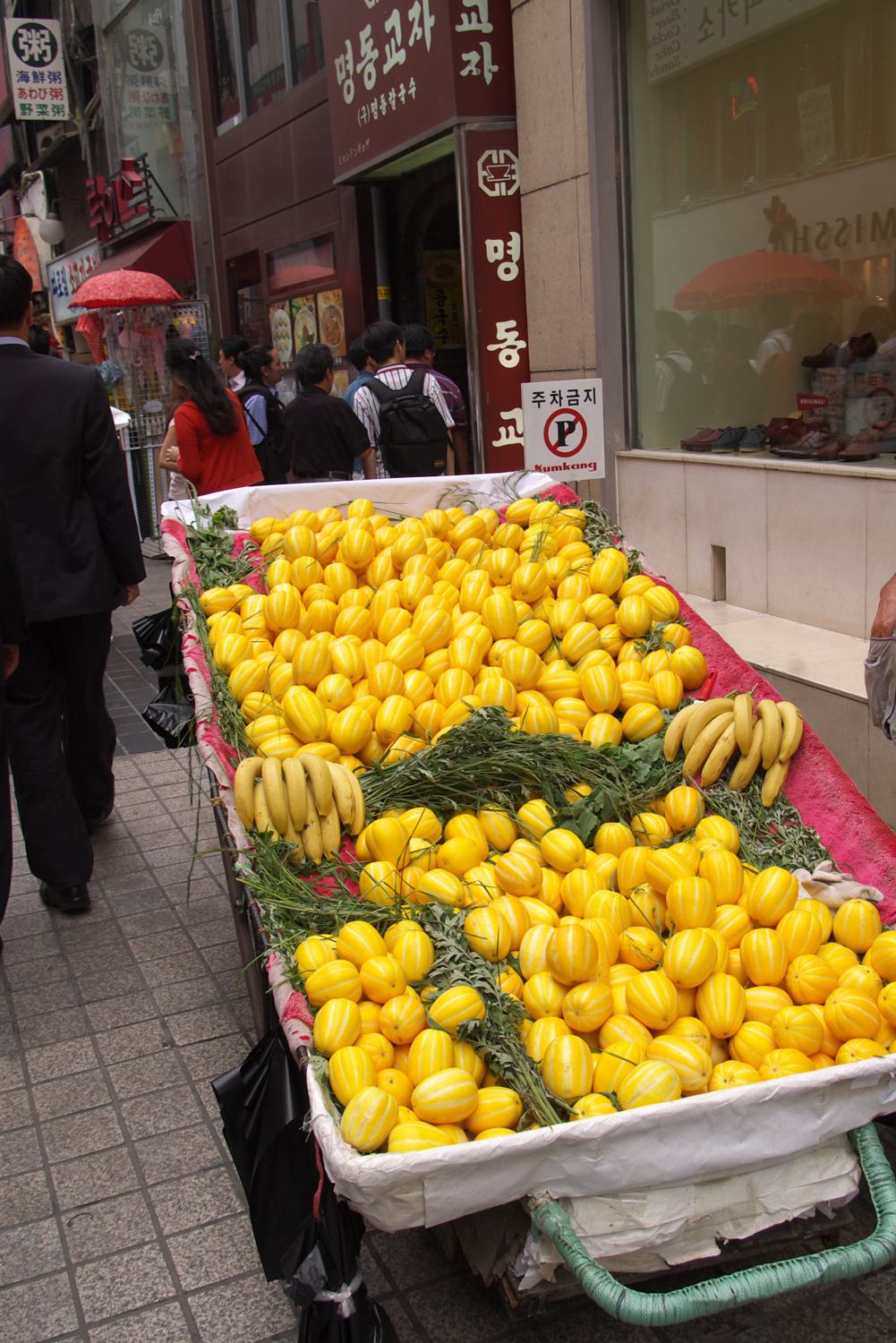A street cart in Seoul piled with yellow chamoe melons, a woman selling from it in front of shopfronts.