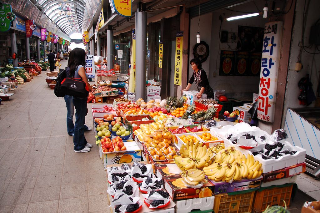 A fruit shop at Seongdong Market in Gyeongju, its front stacked with boxes of persimmons, apples, grapes, bananas and pineapples under a striped awning.