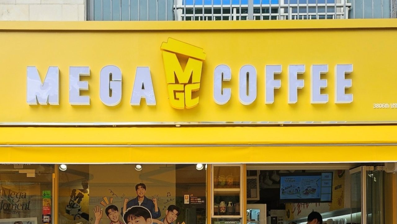 The yellow storefront of a Mega MGC Coffee shop, the sign reading MEGA MGC COFFEE, posters in the window.