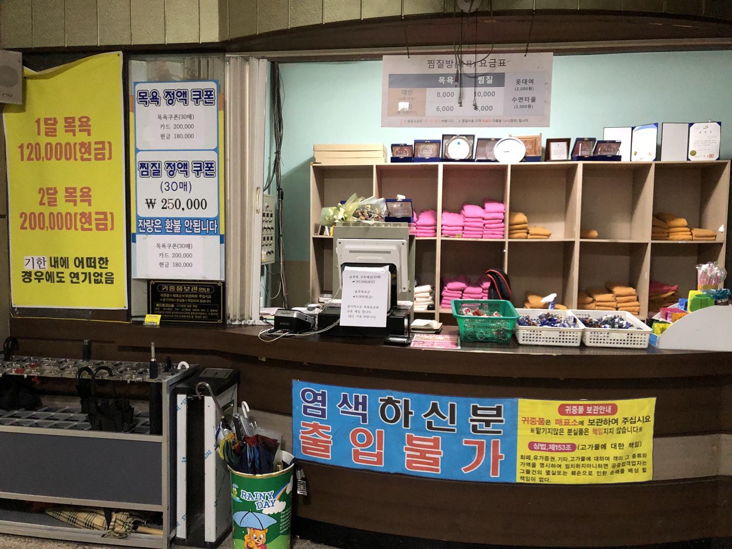 The front counter of a Korean bathhouse in 2020: shelves of folded towels and sauna clothes, a rack of scrub mitts, and yellow signs offering a month of baths for ₩120,000 cash.