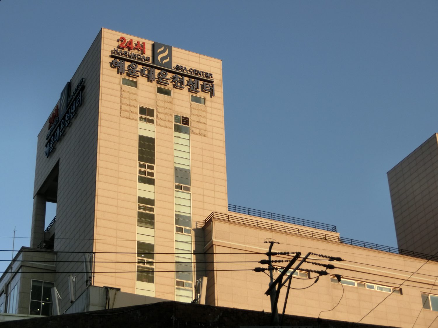 The Haeundae Spa Center in Busan at dusk, a plain concrete bathhouse block with a red 24-hour sign and the name in Korean on the roof.