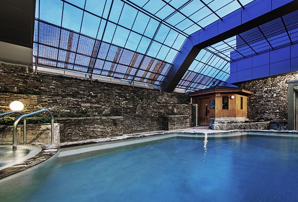 A large indoor hot-spring pool under a glass roof, stone walls and a small wooden sauna hut at the far end, no bathers.
