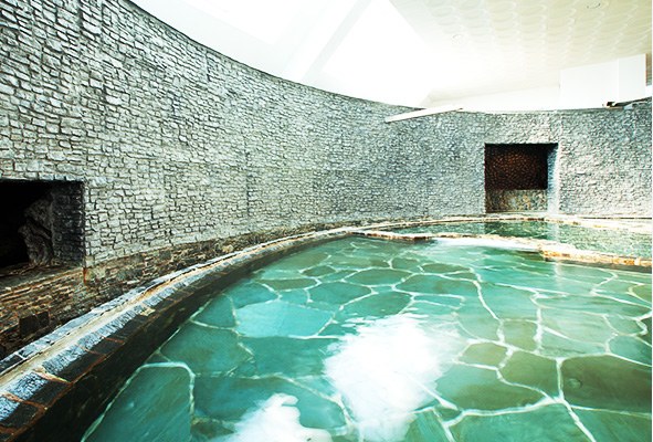 An empty Korean hot-spring bath hall: a curved stone wall, a wide pool of pale green water with a stone floor, no one in it.