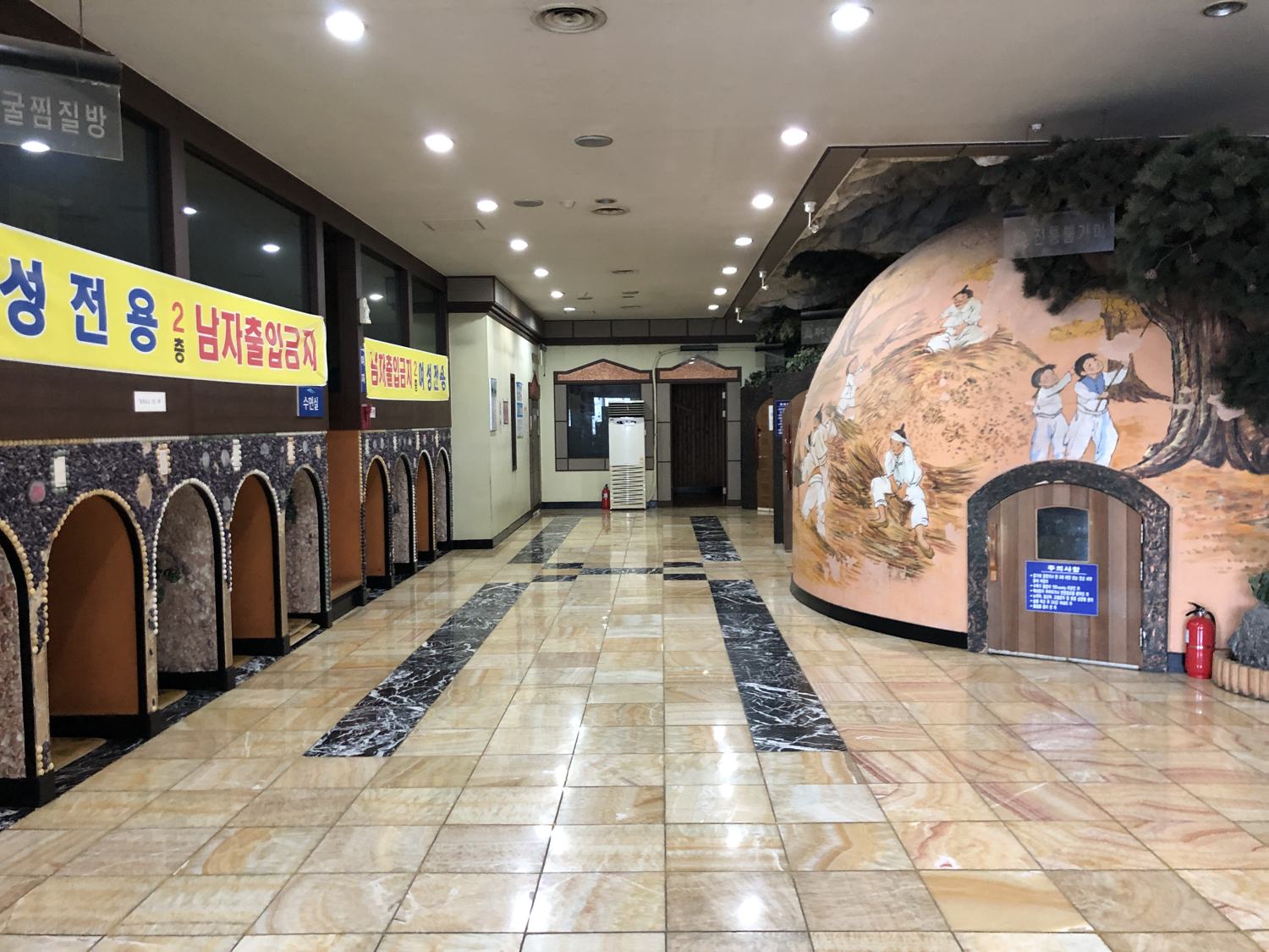 The main hall of a 24-hour sauna in Wonju: a polished stone floor, a mural of a village scene around a domed kiln room, and a banner reading men-only for the second floor.