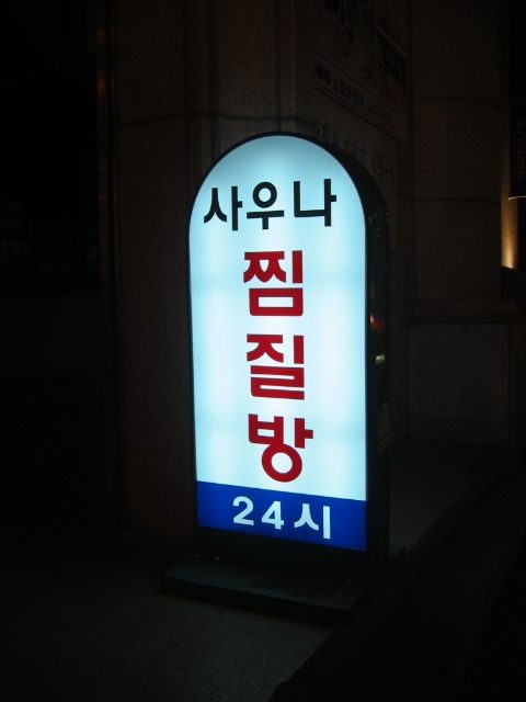 A lightbox sign at night reading, in Korean, sauna, jjimjilbang, 24 hours.