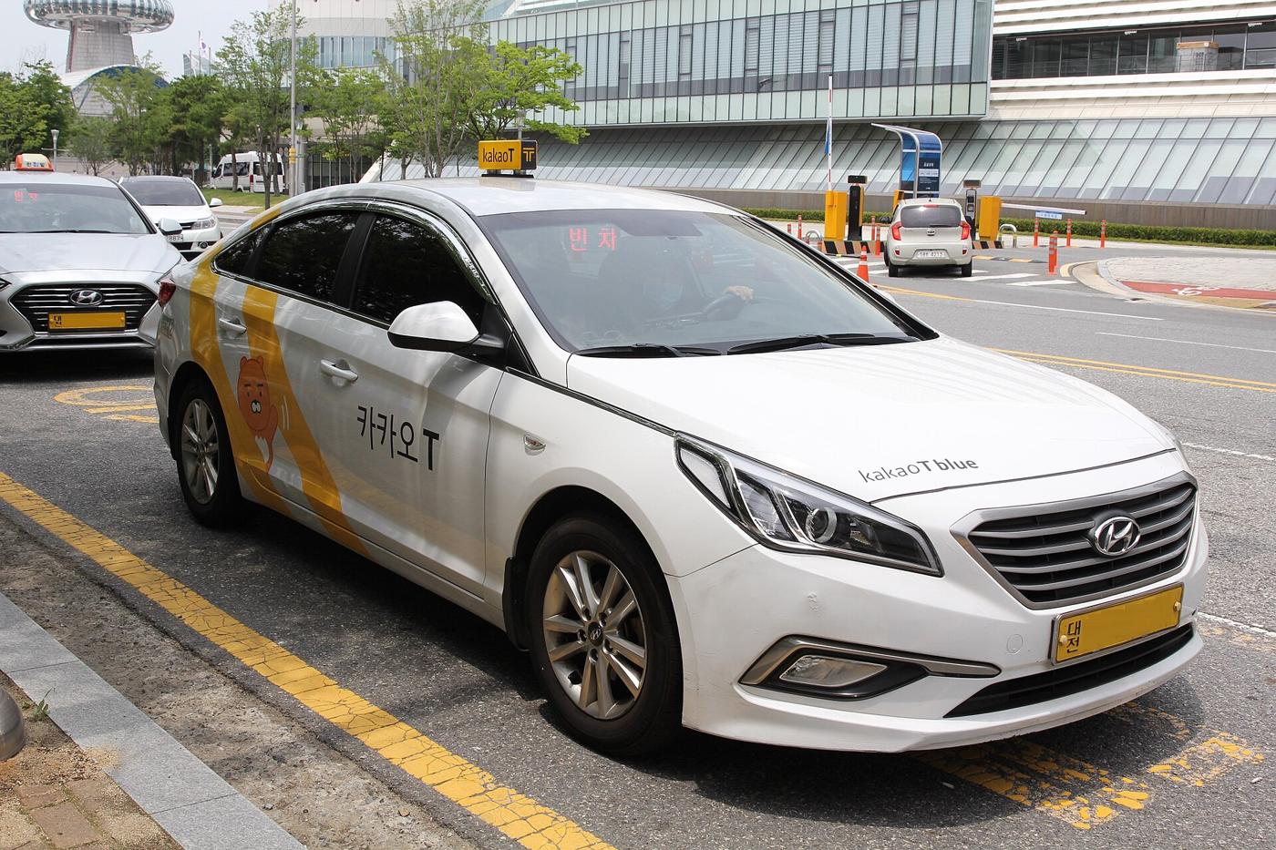 A white Hyundai Sonata taxi in Kakao T Blue livery, roof light reading kakaoT, the windscreen sign showing 빈차 for vacant.