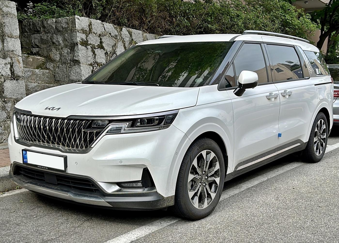 A white Kia Carnival minivan parked beside a stone wall on a Seoul street.
