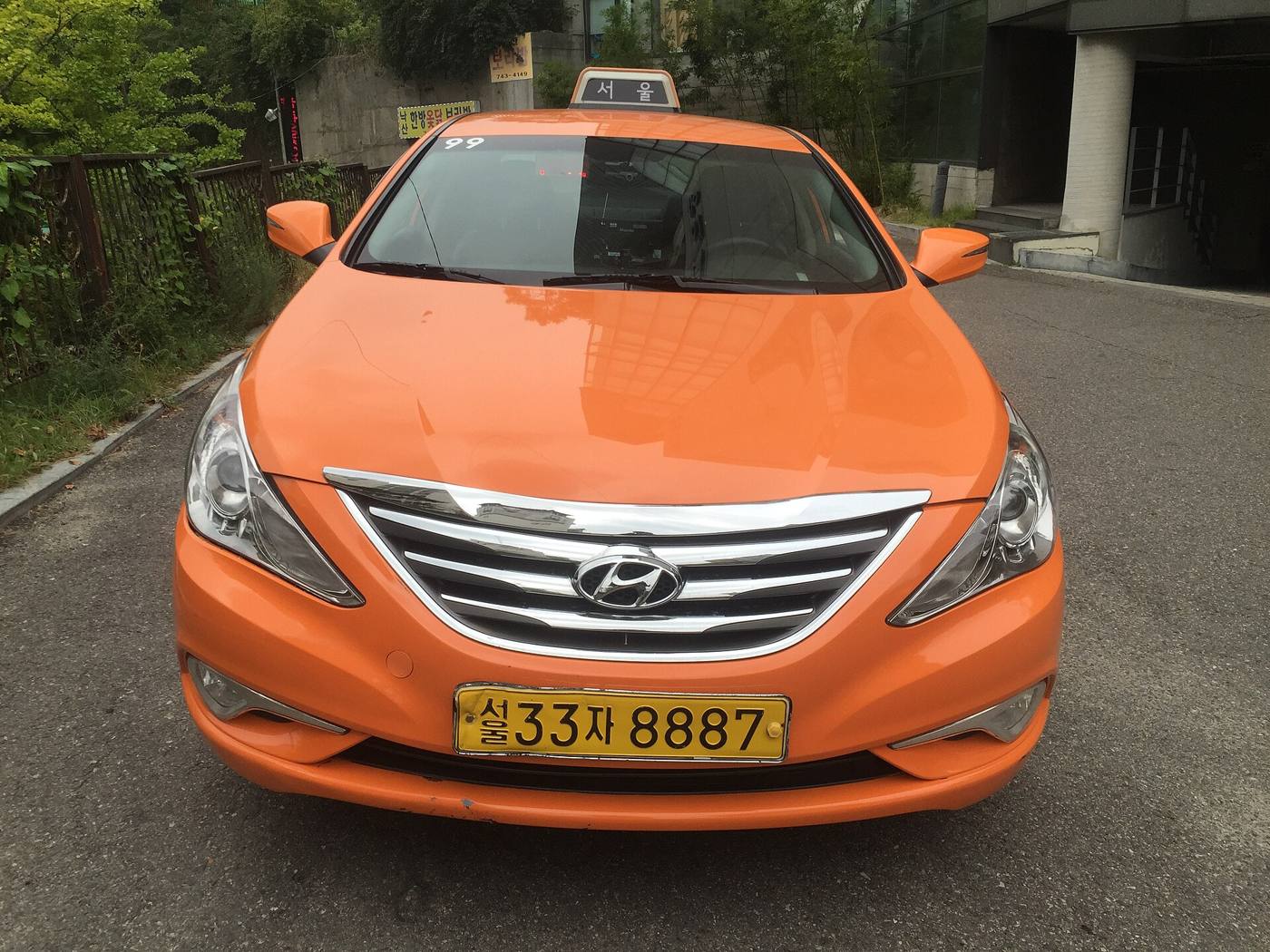 An orange Seoul sedan taxi, a Hyundai Sonata with the 서울 roof sign and yellow plate, parked on a hillside street.