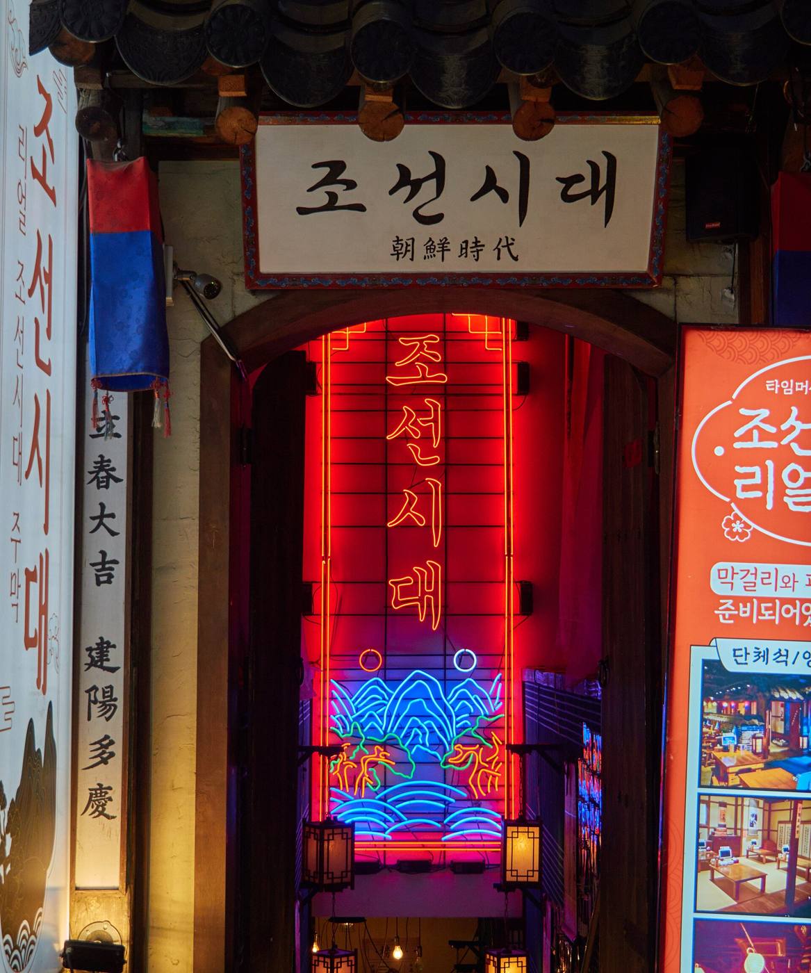 The entrance of Joseon Sidae, a traditional-themed restaurant and bar in Hongdae, Seoul, with a tiled roof, a calligraphy signboard and red neon lettering inside the doorway.
